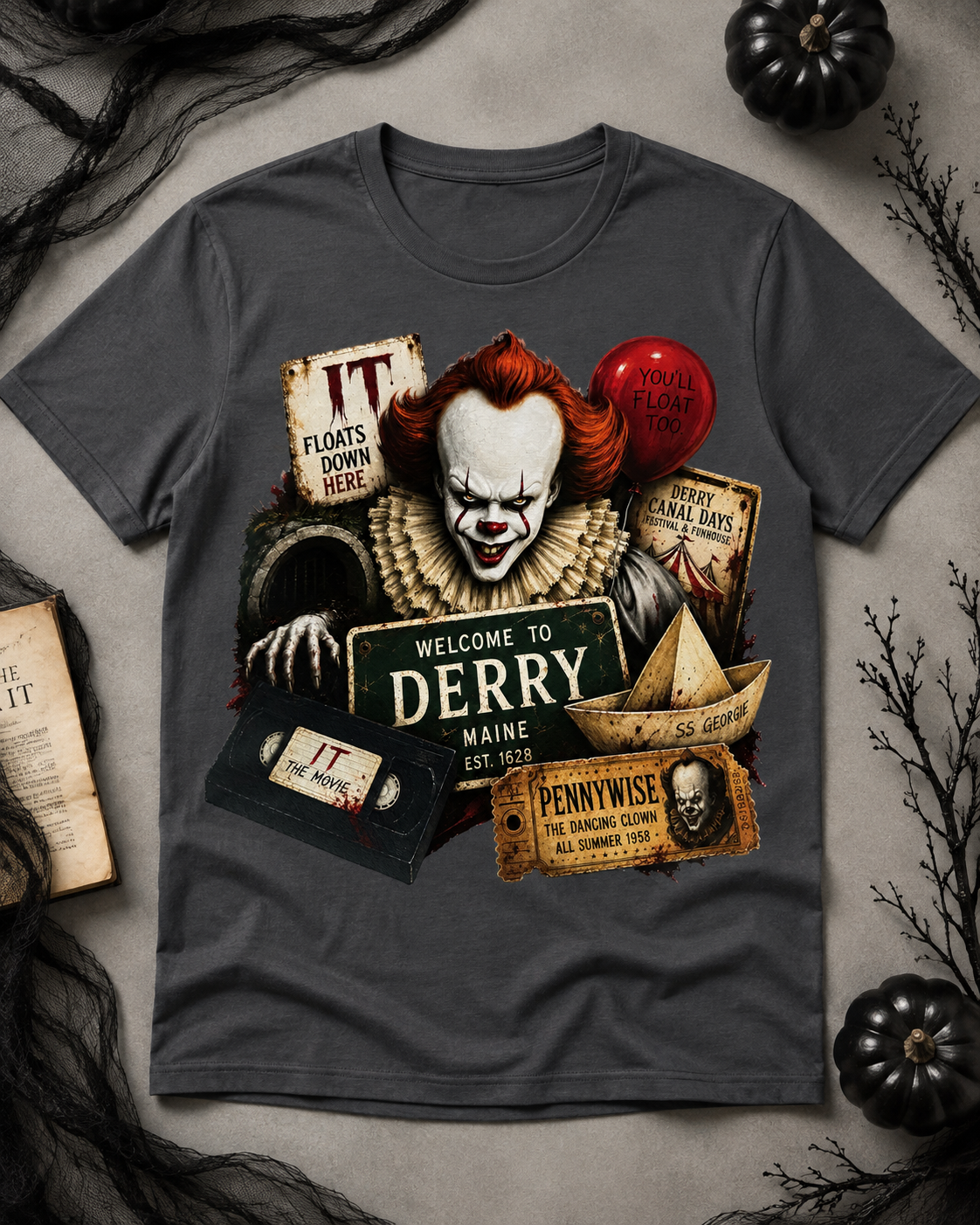 A dark gray graphic t-shirt featuring a Pennywise the Clown character design from the movie IT, styled in a flat-lay with Halloween props like black spiderwebs, dried branches, and small black pumpkins on a textured gray background.